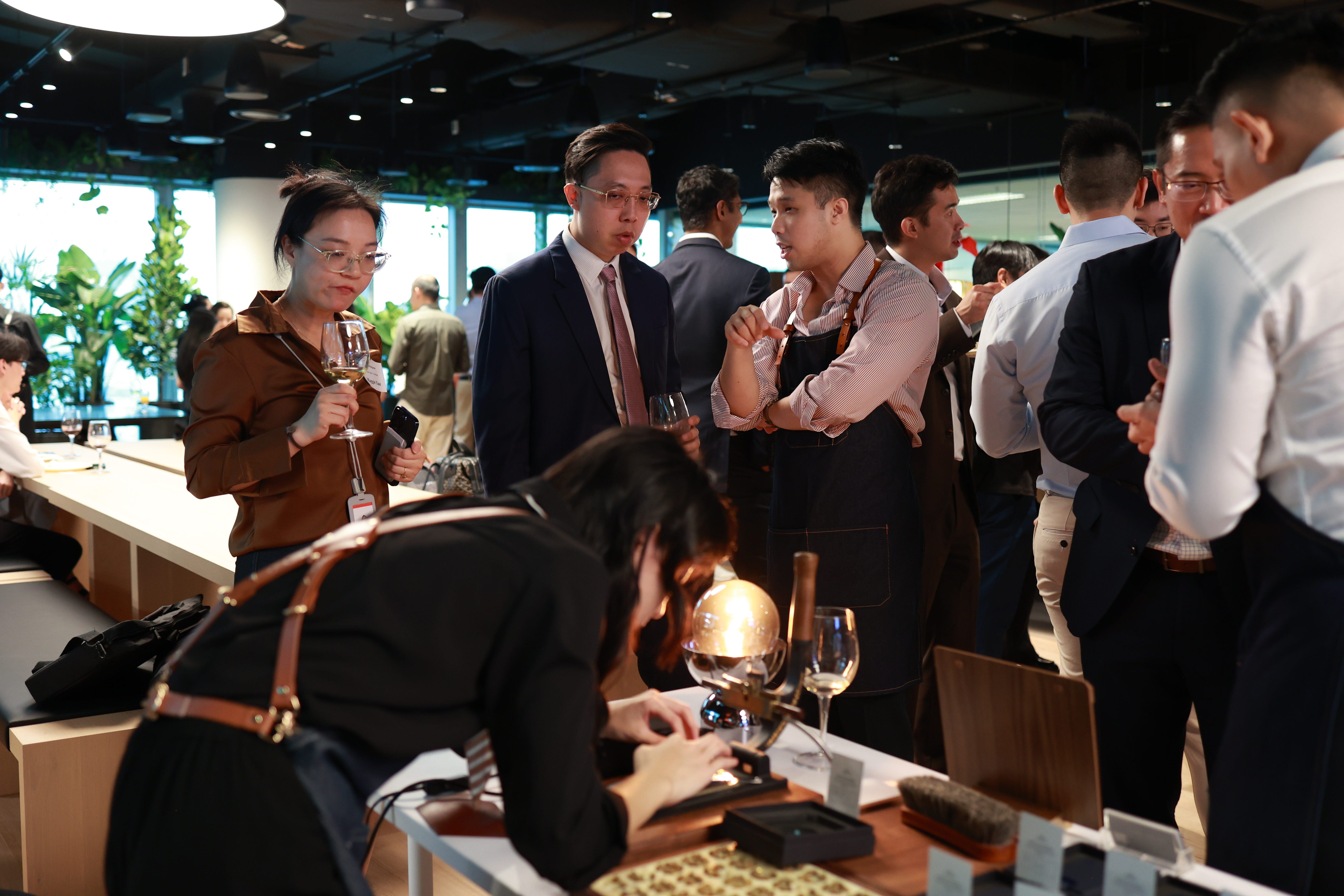 A busy indoor networking or event scene with multiple people standing and interacting around a table while Dechen is performing personalisation of the leather corporate gift. Several men in business attire are conversing, while one person in the foreground leans over the table.