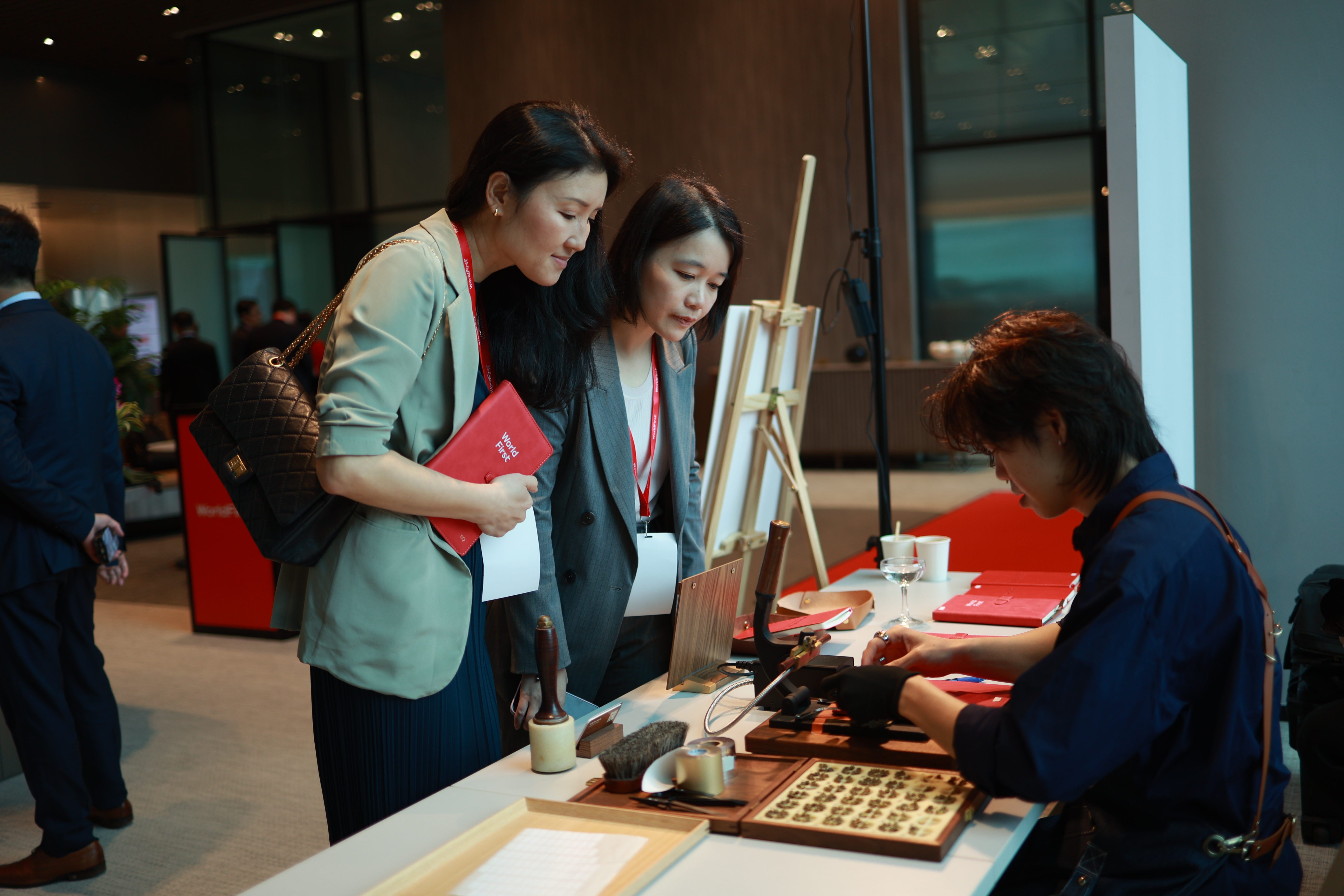 A lifestyle scene at a traditional Chinese calligraphy workshop or exhibition, showing three women observing a calligraphy demonstration by a seated artisan at a table working to personalise leather with embossing machine