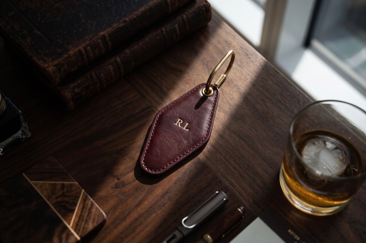 A dark reddish-brown vintage leather key fob with brass hardware and an engraved 'RL' monogram, resting on a dark wooden table next to a glass of whiskey and books.