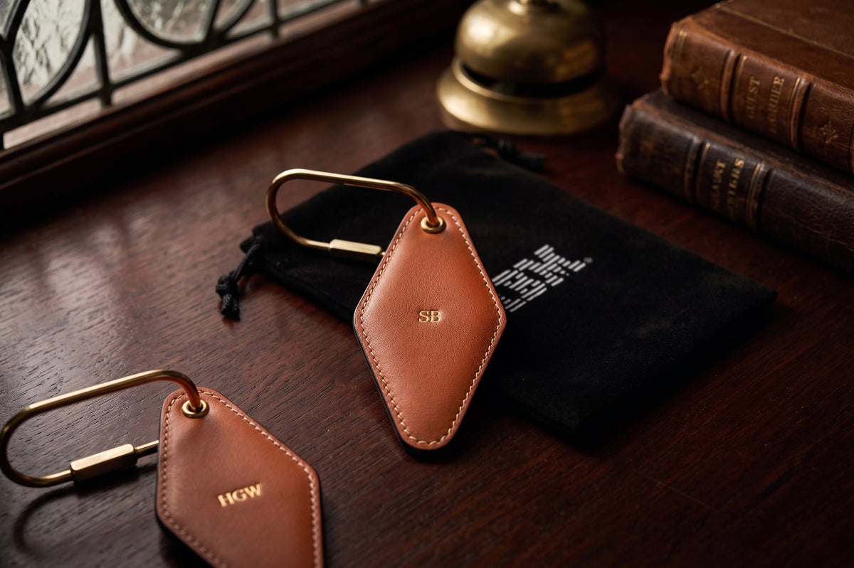 Two tan vintage hotel leather tags with gold-tone solid brass metal rings, displayed on a dark wooden table next to a black drawstring pouch, with vintage books and a brass bell in the background.