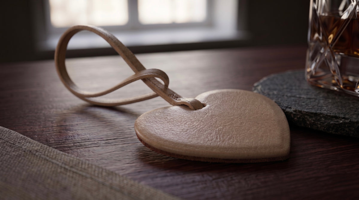 A heart-shaped beige leather bag charm with a matching black leather loop, resting on a dark wooden table next to a glass of whiskey.