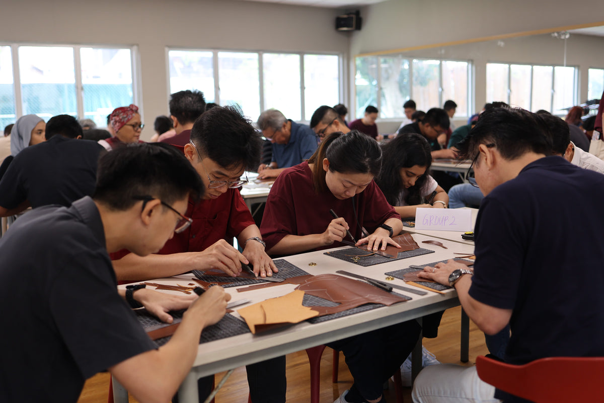 A group of people seated at long tables in a bright workshop, crafting leather products. Large windows line the back wall, and several participants are visible in the background.
