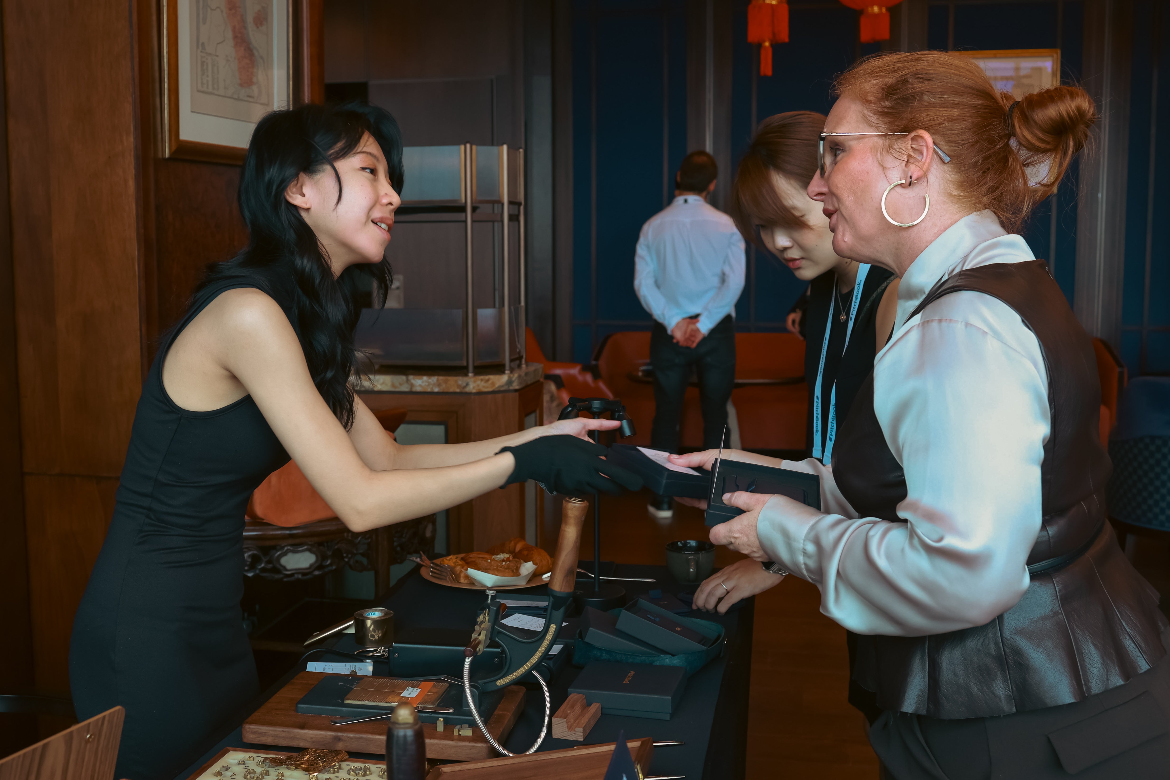 A woman in a black sleeveless dress, and black gloves - craftsman from The Leather Society extends the completed embossed with initials Navy Magsafe Cardholder another woman wearing a white blouse, dark vest, and glasses in an indoor setting with other people and tables visible.