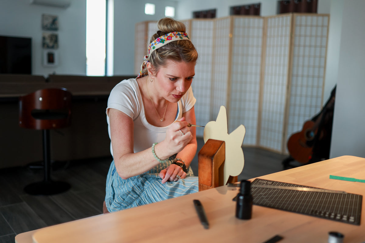 A woman in a white t-shirt and blue skirt sits at a wooden table, carefully painting a calvi inspired cardholder with an awl. The table has a black marker and a small black bottle, and a guitar is visible in the background of the room.
