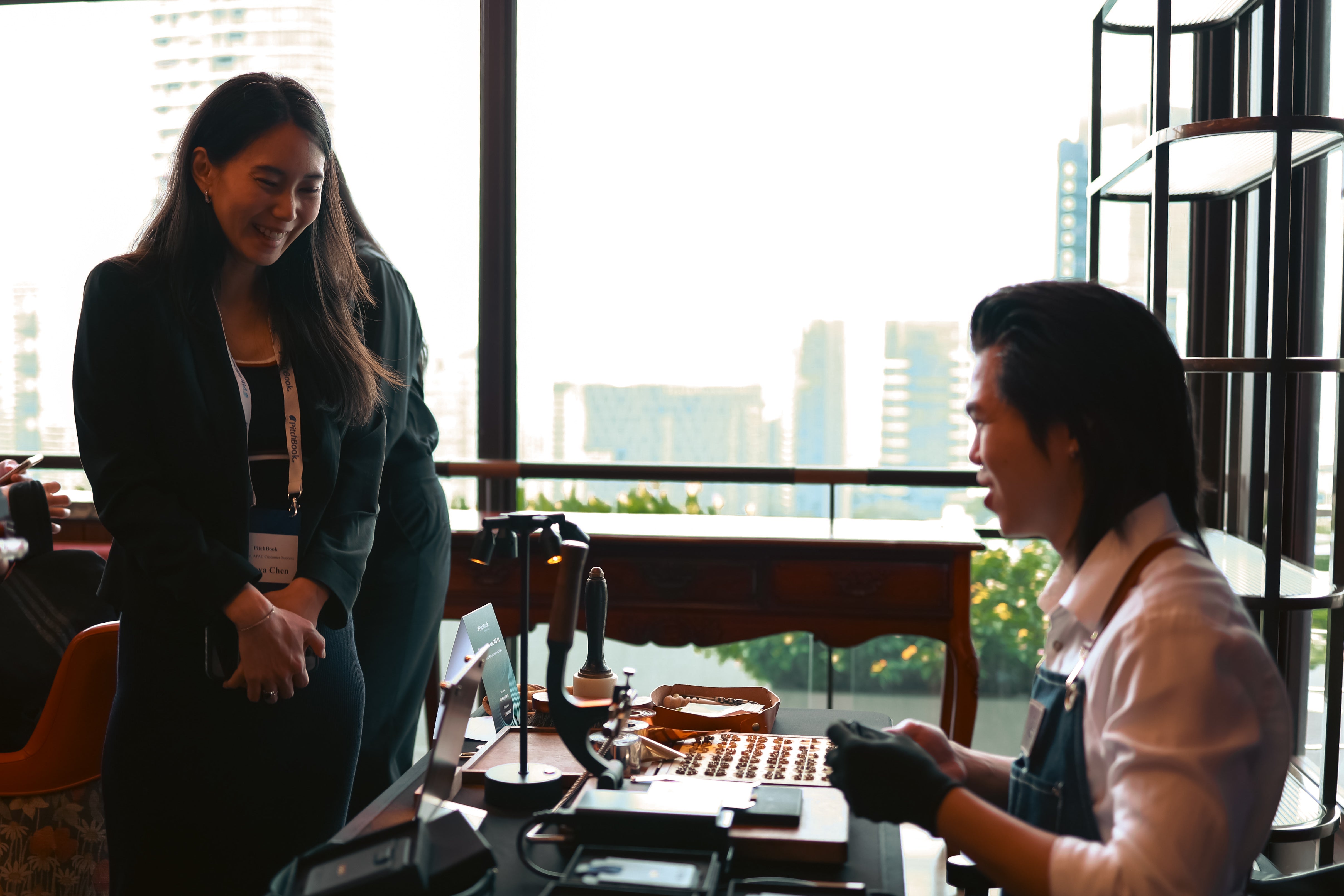A woman in a black blazer stands smiling while talking to a The Leather Society craftsman wearing an apron and gloves behind a table displaying magsafe leather wallet about to be embossed, set against a bright window view of a city skyline.