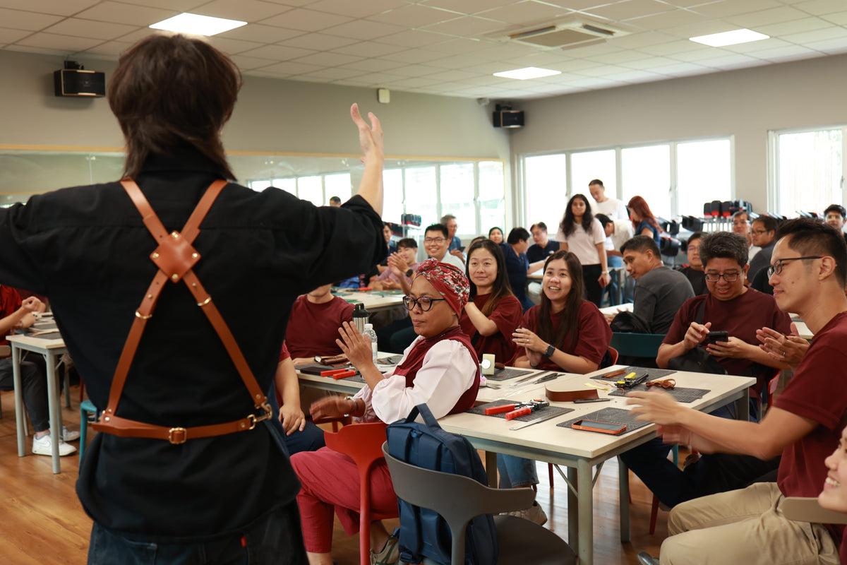 A person in a black shirt with brown leather harness-style straps stands with one arm raised in a workshop setting, facing a seated audience of people at tables explaining more about leather crafting to them.