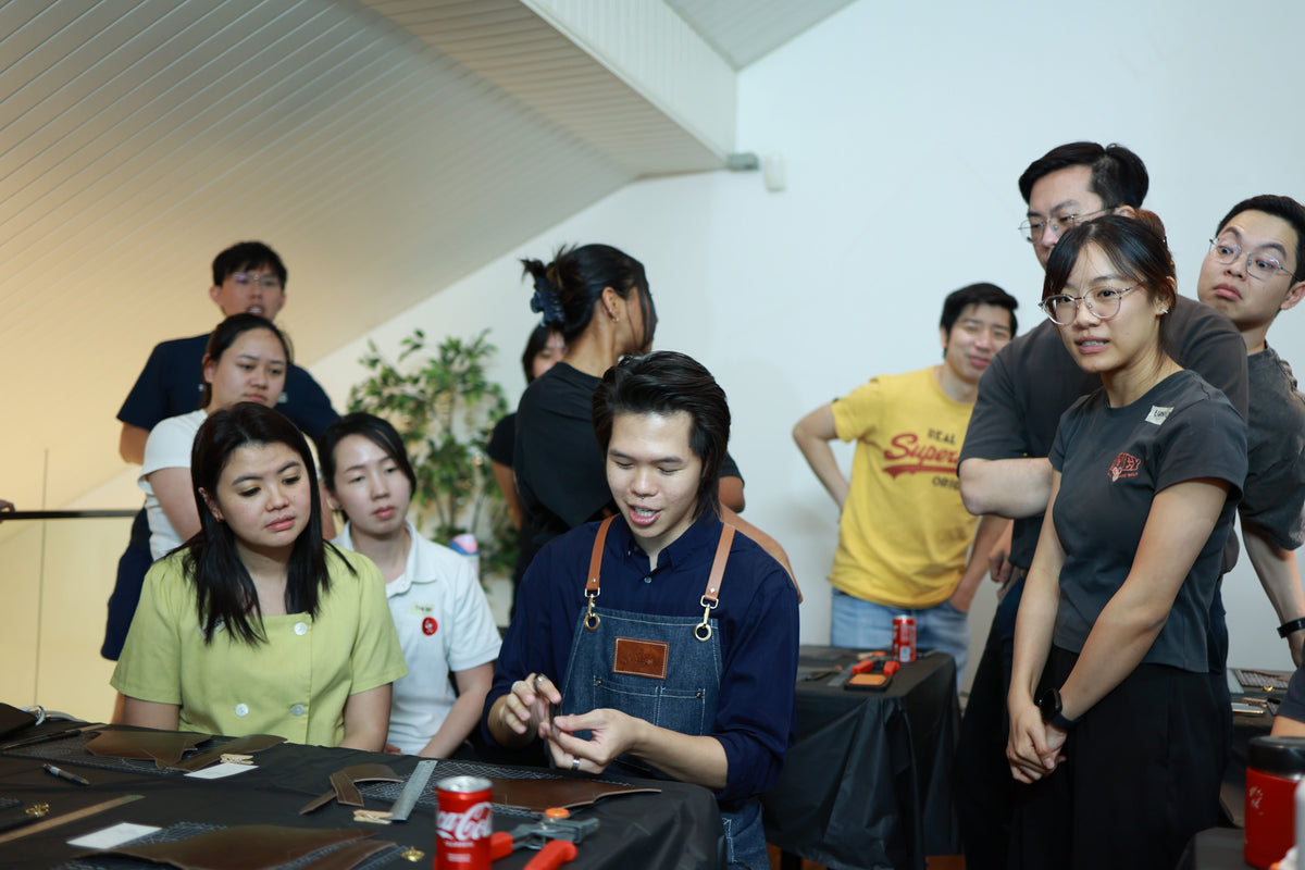 A group of people gathered around a table in a workshop or classroom setting, with instructor keith  in the center wearing a blue apron and holding a small object teaching and explaining more about leather crafting technique, while others watch or observe. The scene is indoors with white walls, ceiling panels, and a plant in the background.