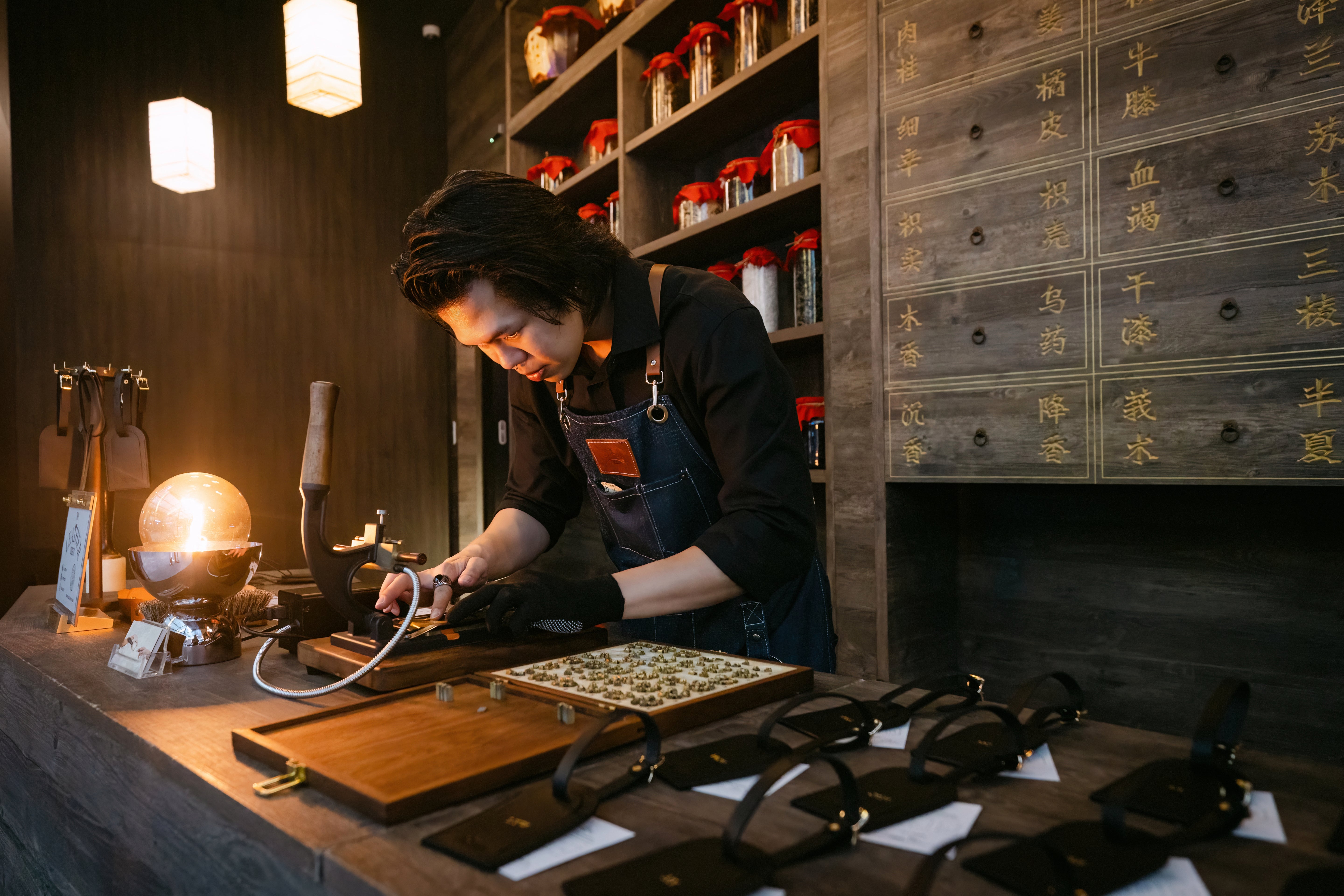A craftsman wearing a dark apron and gloves works at a wooden table inside a dimly lit shop, embossing a leather piece. The counter is cluttered with leather straps, tools, and a glowing lamp, while shelves in the background display numerous glass jars with red lids.