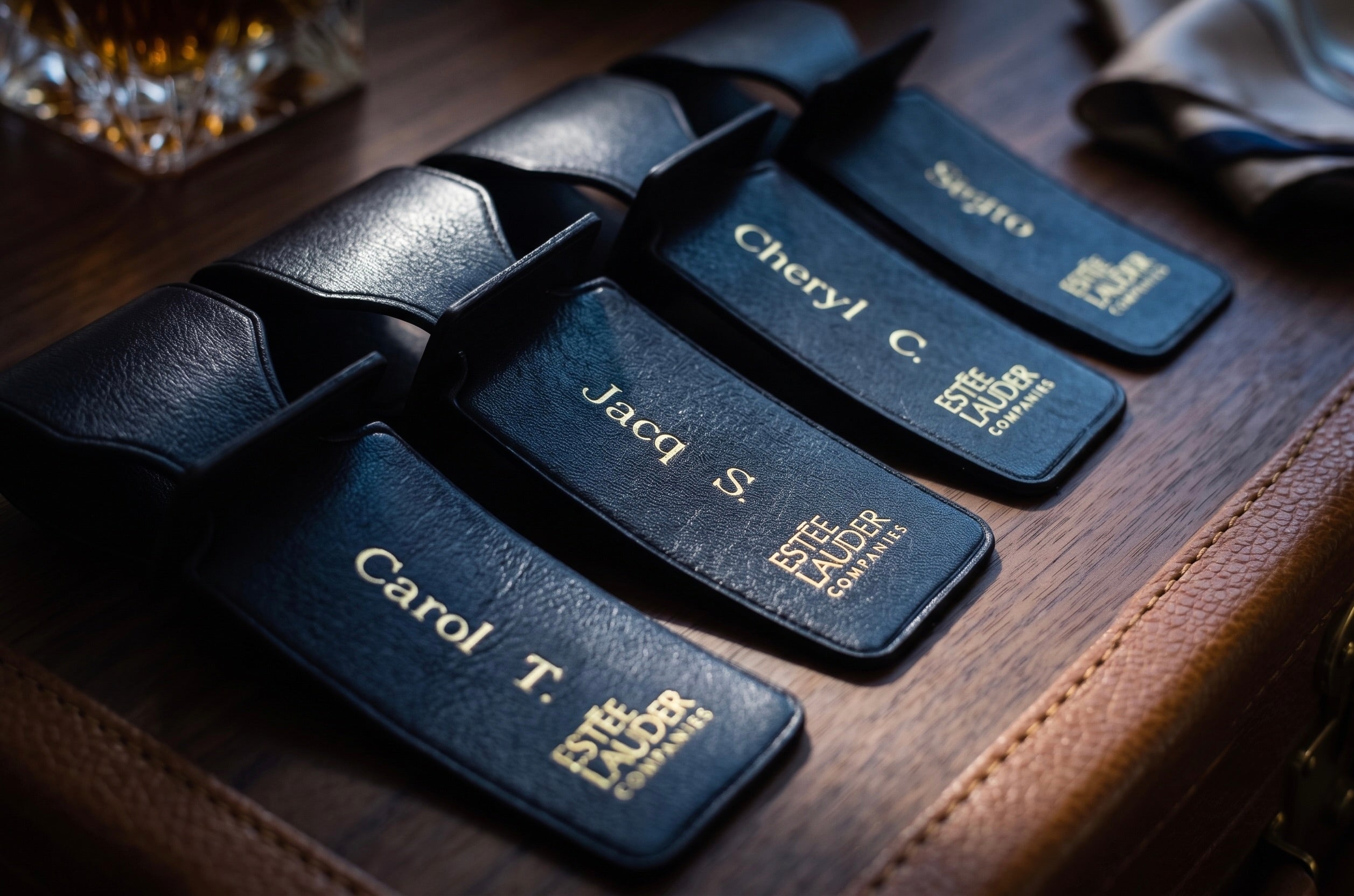 Four black leather luggage tags with gold-embossed names and 'Estee Lauder Cosmetics' logos, arranged on a dark wooden surface.