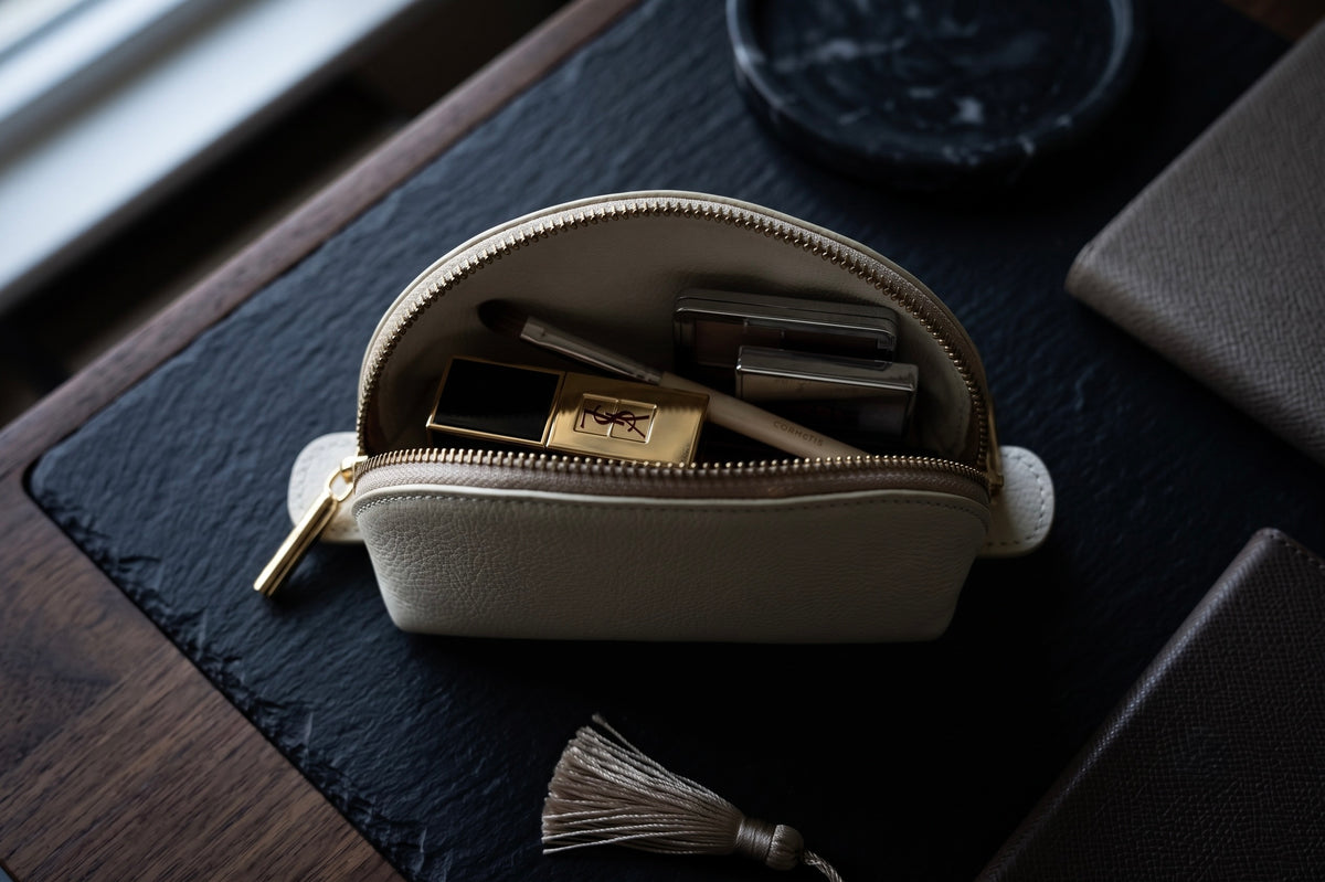 A cream-colored leather cosmetic pouch with a gold zipper and tassel, shown open and filled with makeup items on a dark textured mat.