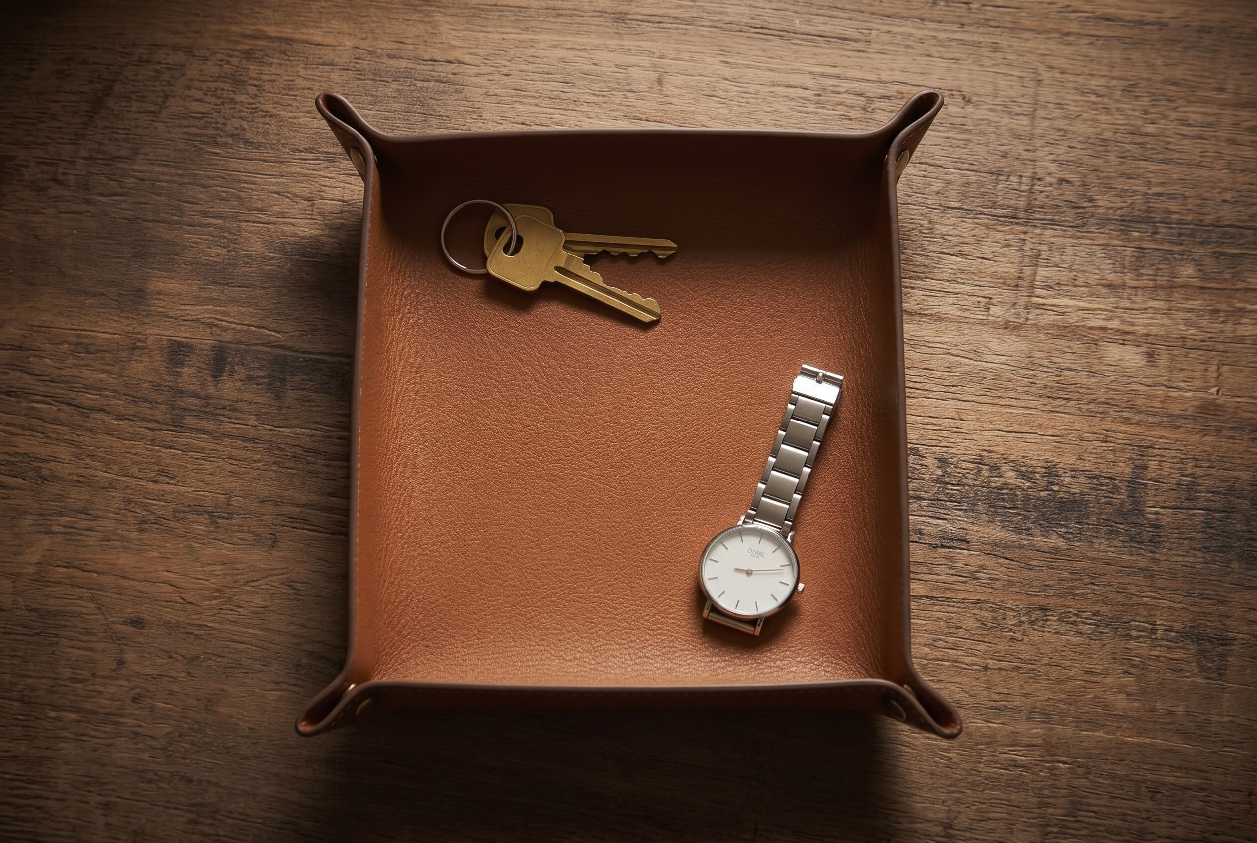 A square brown leather valet tray with folded corners, shown on a dark wood tabletop. Inside the tray are a set of gold keys and a silver wristwatch with a white dial.