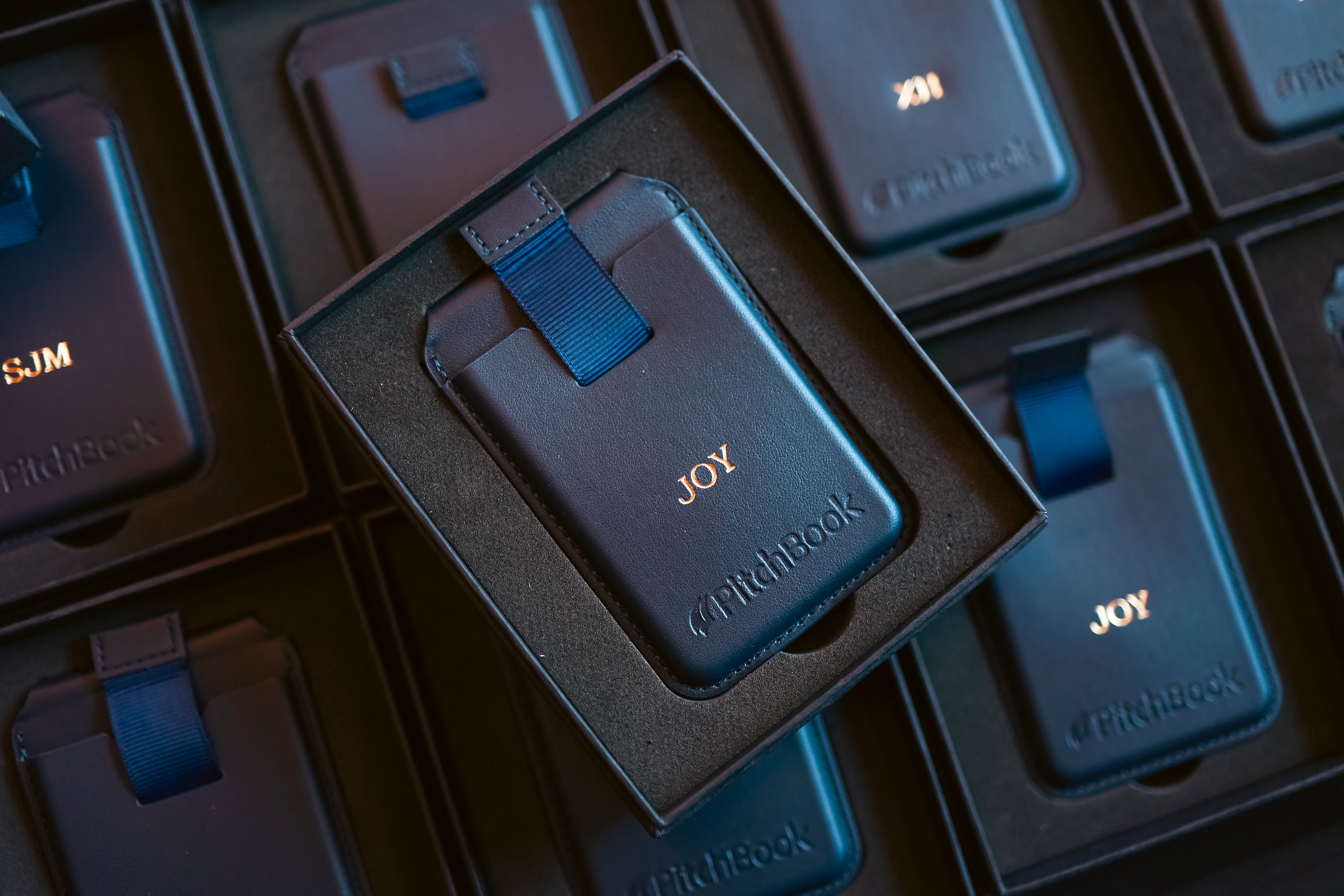 A close-up of a dark blue FitchBook card magesafe cardholder with a blue strap, displayed inside an open black presentation box. The card holder is embossed with the brand name 'FitchBook' and the word 'JOY'.