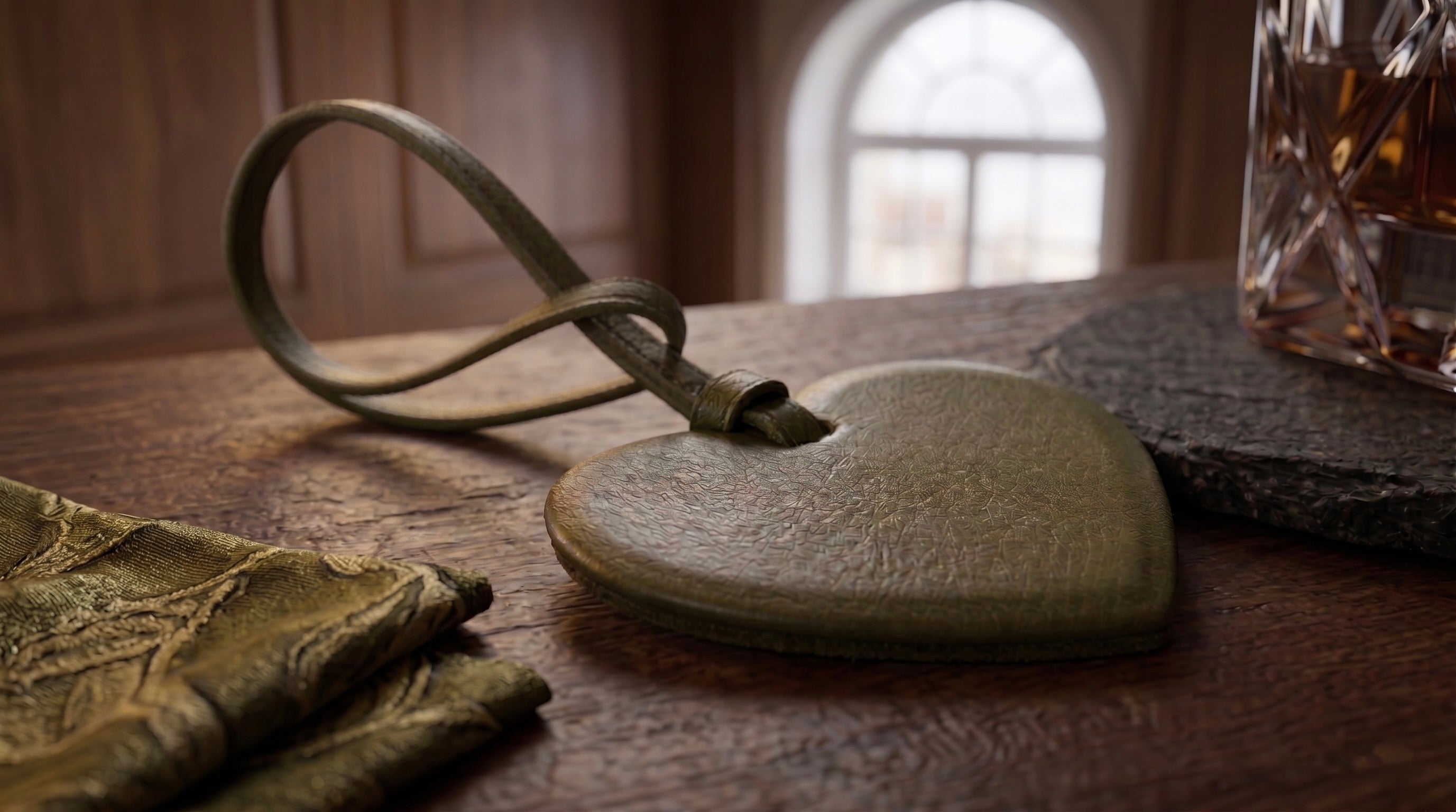 A heart-shaped olive leather bag charm with a matching olive leather loop, resting on a dark wooden table next to a glass of whiskey and high end scarf.