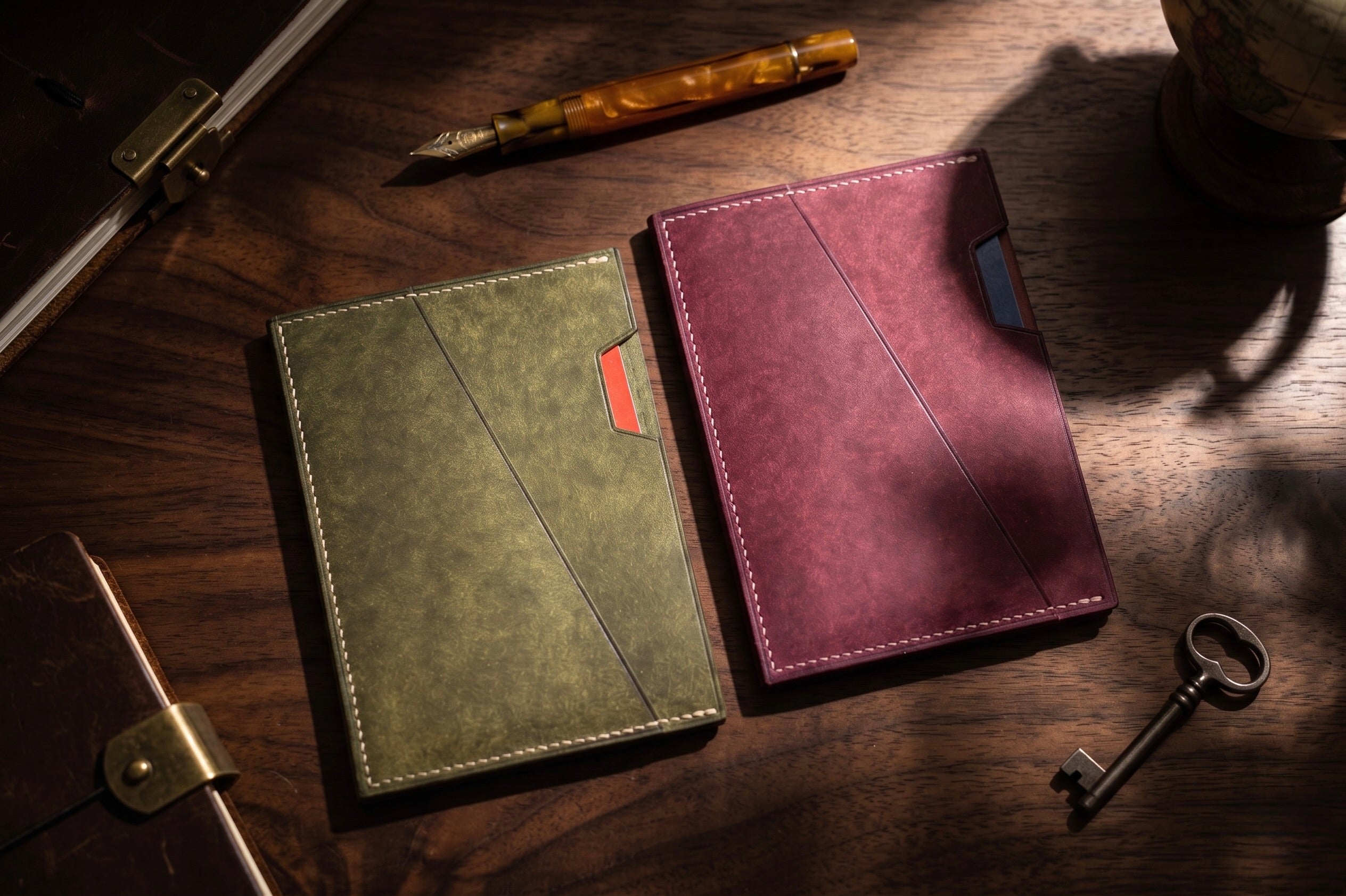 Two leather passport sleeves on a wooden desk, one in olive green pueblo with a singapore passport and one in burgundy with a united states passport. A fountain pen, key, and other desk props are visible around them.