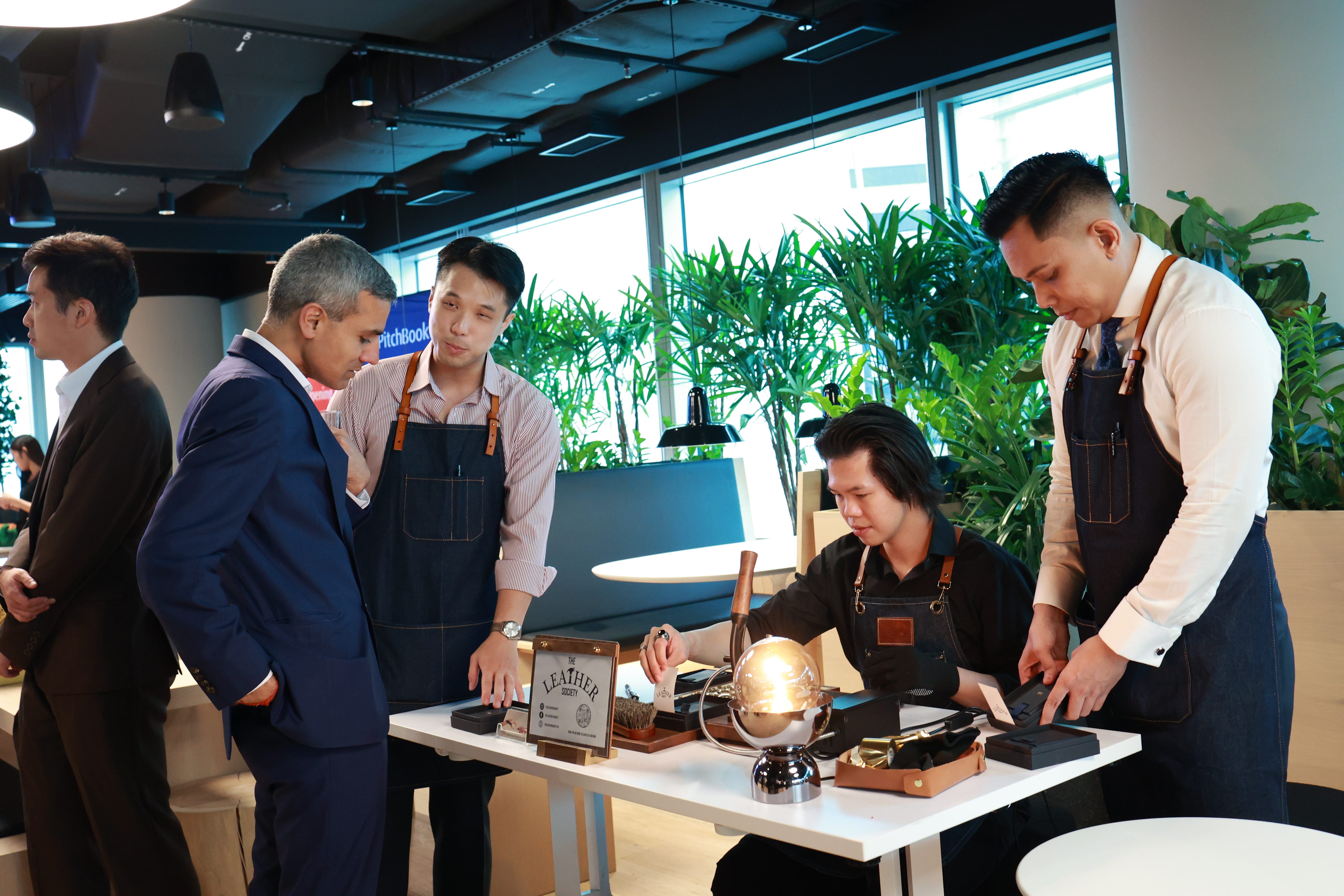 CEO of Pitchbook and and group of men in aprons gathered around a table displaying leatehr embossing machine and instruments in a modern office setting with large windows and green plants.