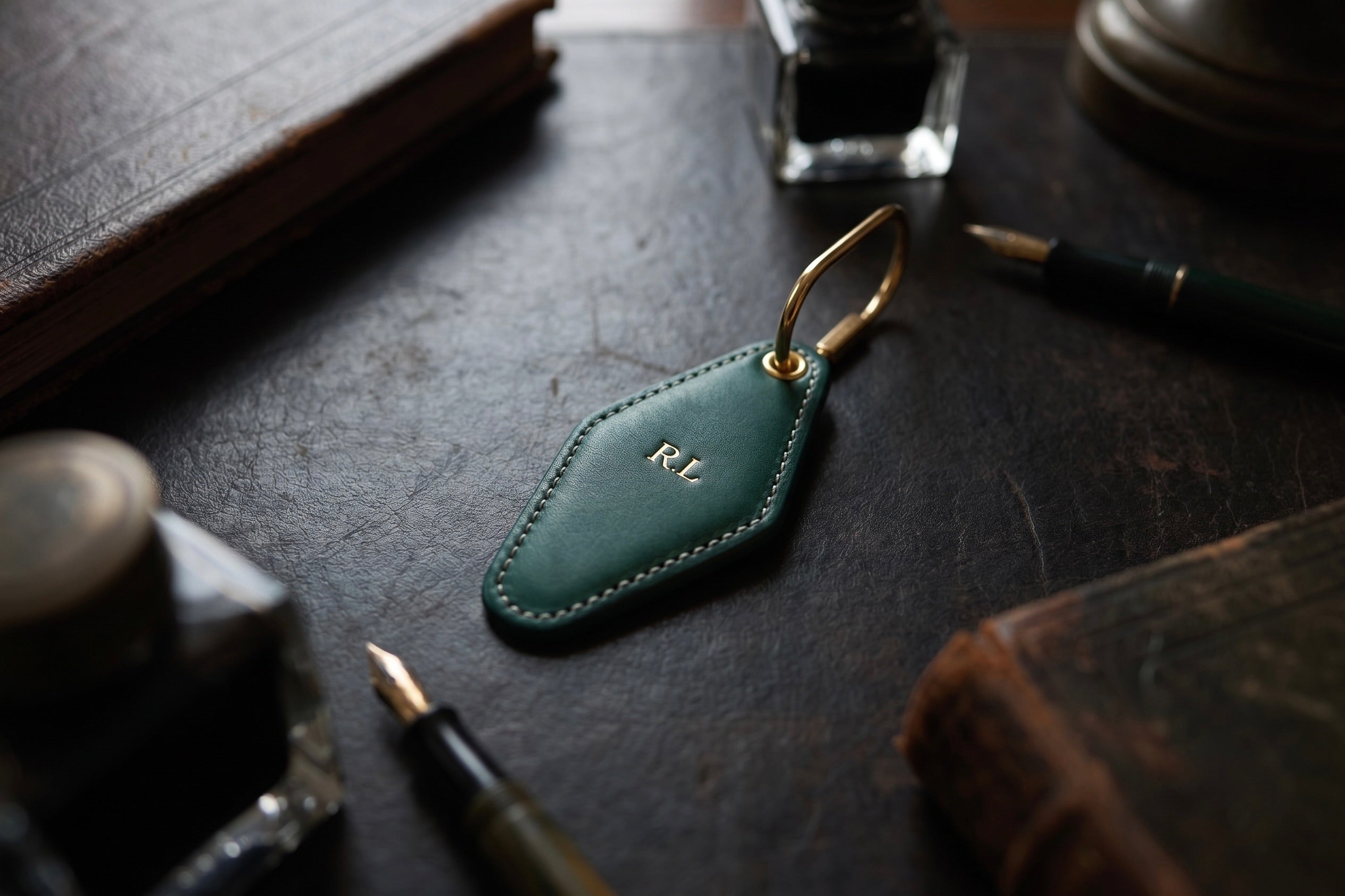 A dark green Vintage Leather Hotel Tag with gold-tone brass hardware and an engraved 'RL' monogram, displayed on a dark desk surrounded by vintage stationery props.