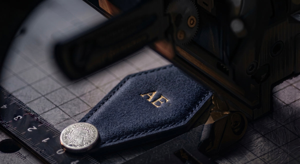 A close-up of a dark navy blue leather monogrammed vintage hotel tag with gold lettering, resting on a metal grid surface next to a magnet and a ruler, with a blurred embossing machine object in the background.